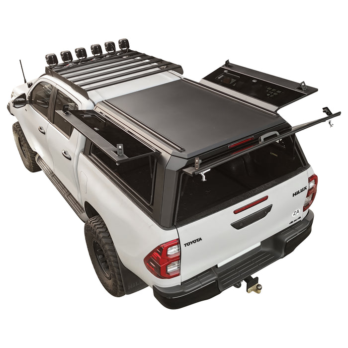 A white Toyota Hilar pickup truck shown from a rear three-quarter angle, fitted with a black hardtop canopy over the bed, and a rear window opening. The product appears to be the truck canopy/accessory.